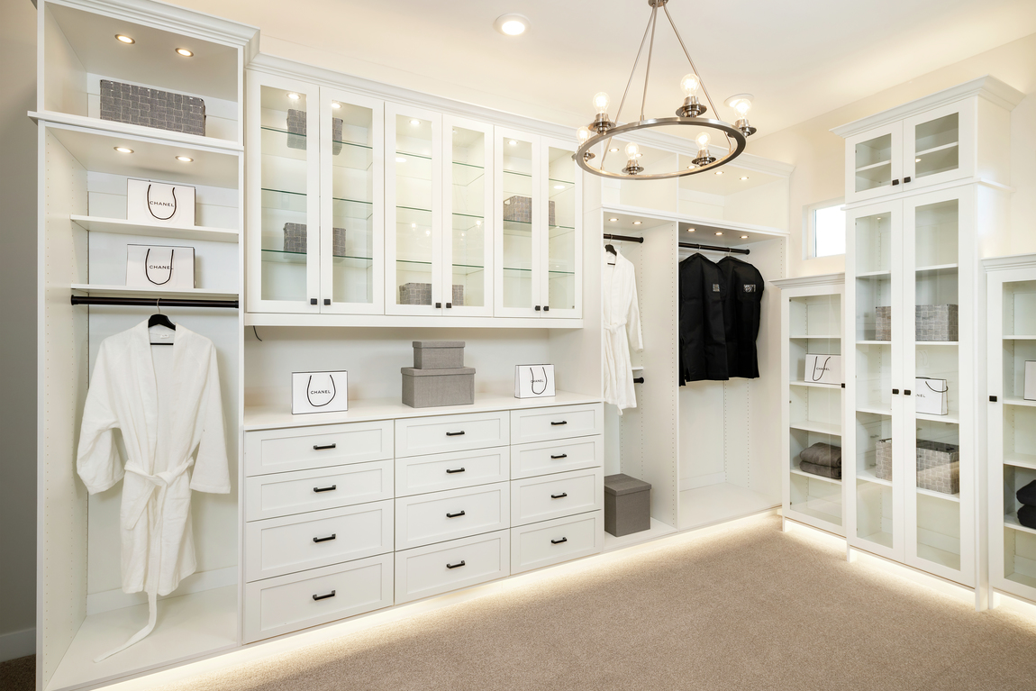 White closet cabinets with toe-kick lighting, spotlights, chandelier and glass cabinet doors.
